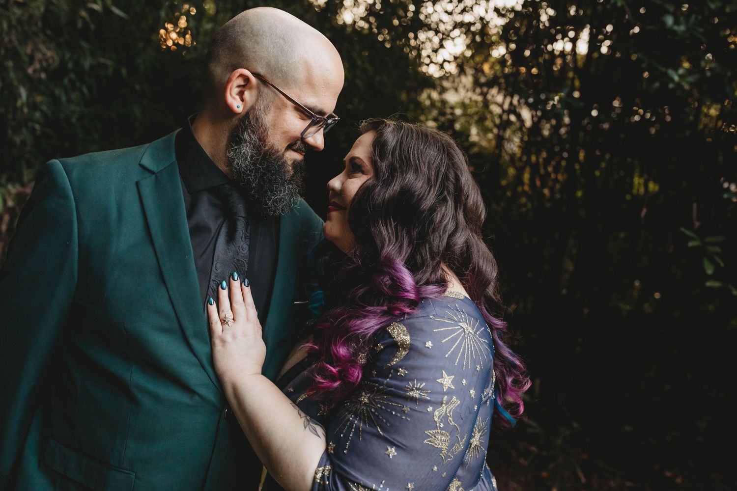 A couple in formal attire gazes lovingly at each other surrounded by lush greenery at sunset.