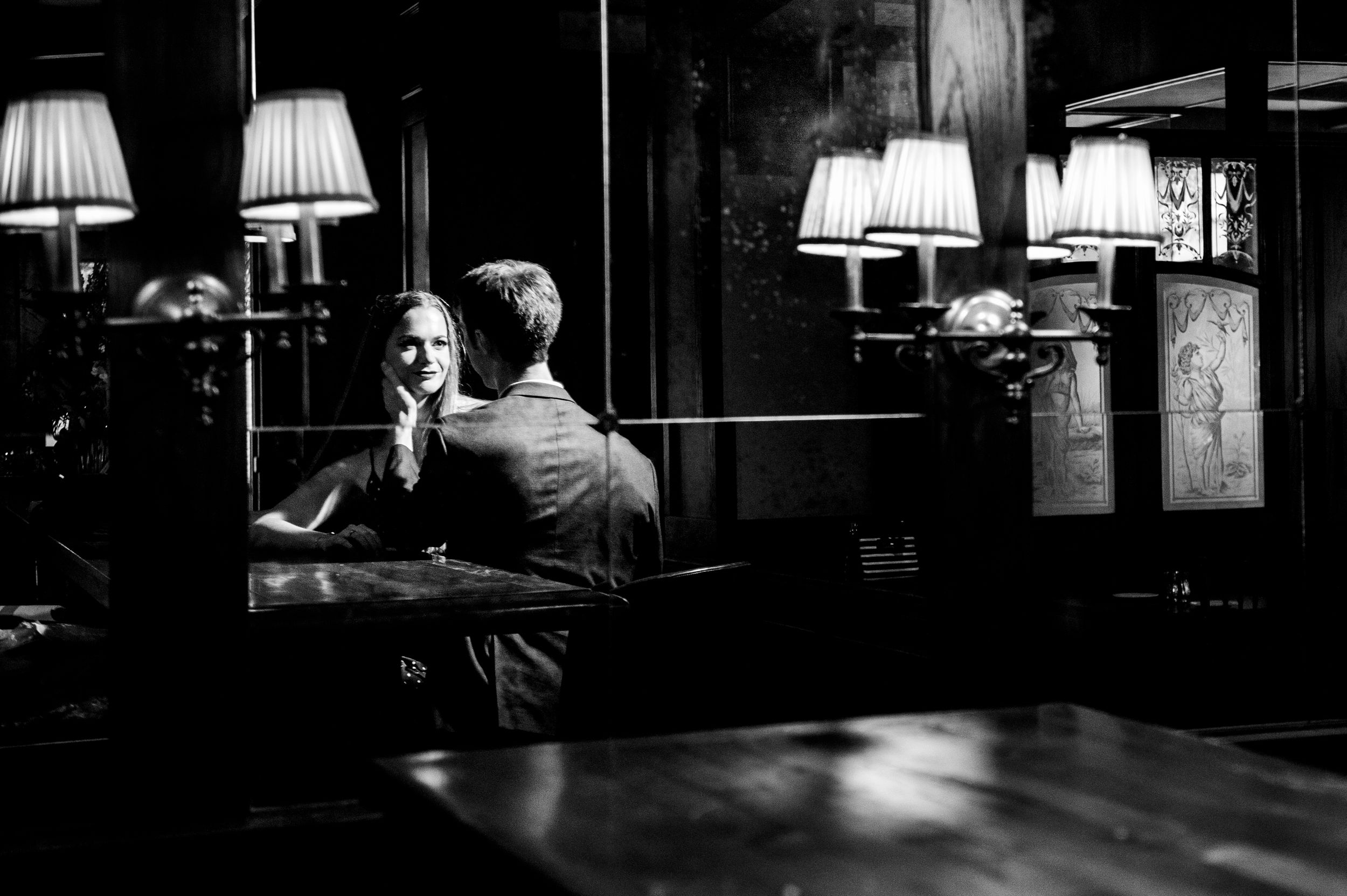 Dramatic black and white noir photograph shows a dimly lit room with table lamps casting moody shadows.