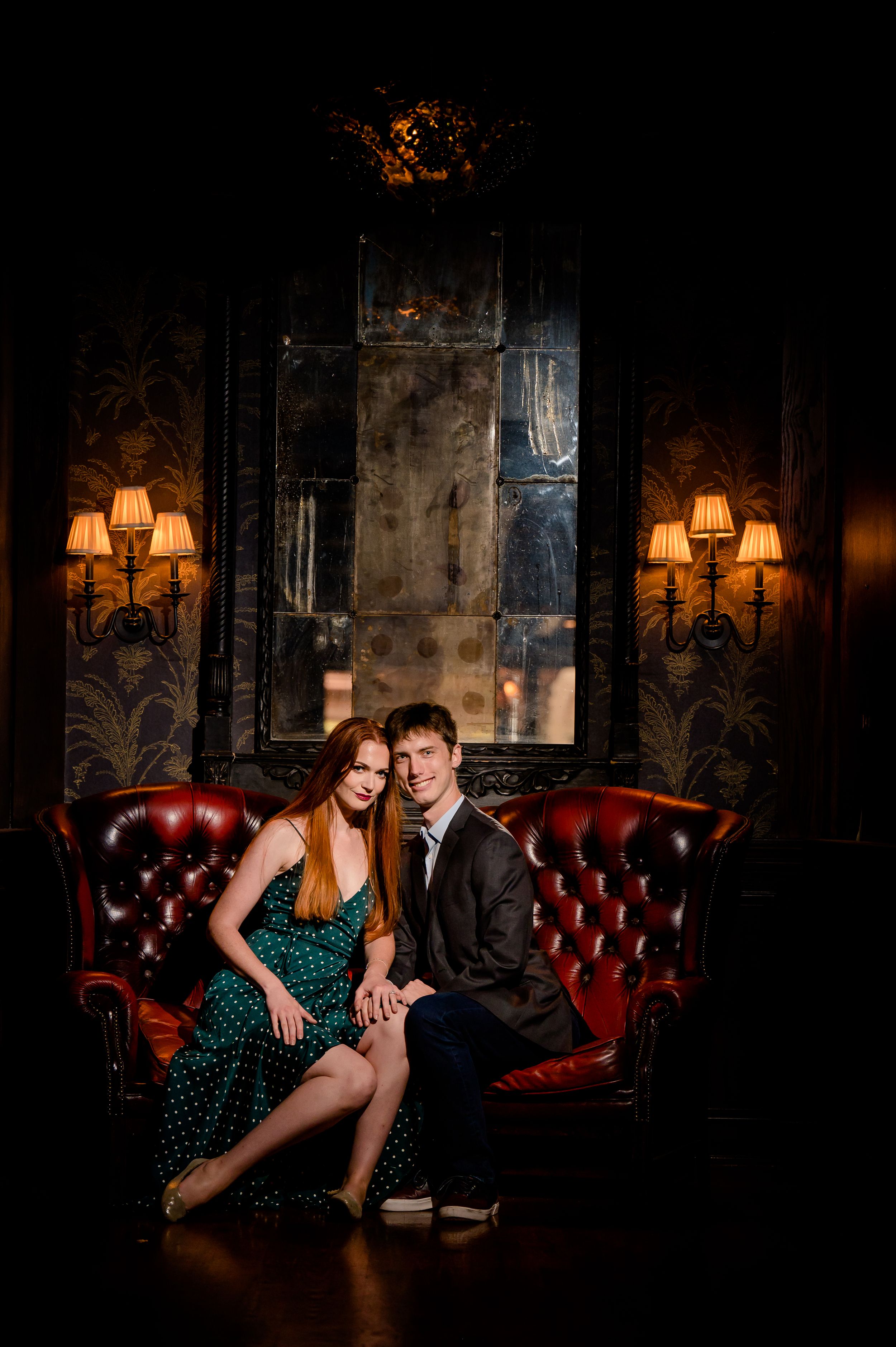 A couple sits together on a vintage red leather Chesterfield sofa in a dark moody room with ornate wall sconces.