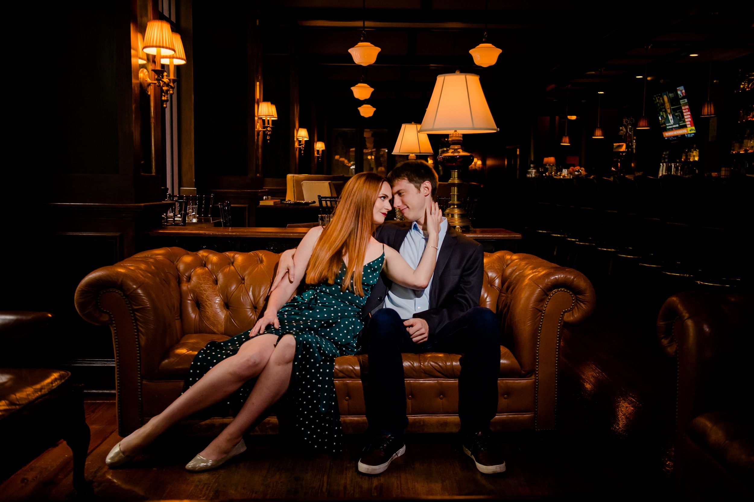 A romantic couple sits on a brown leather sofa in a dimly lit lounge setting with warm ambient lighting.