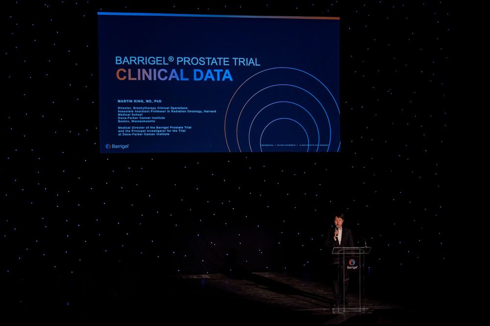 A darkened auditorium stage with a podium and blue clinical data presentation slide projected on screen.