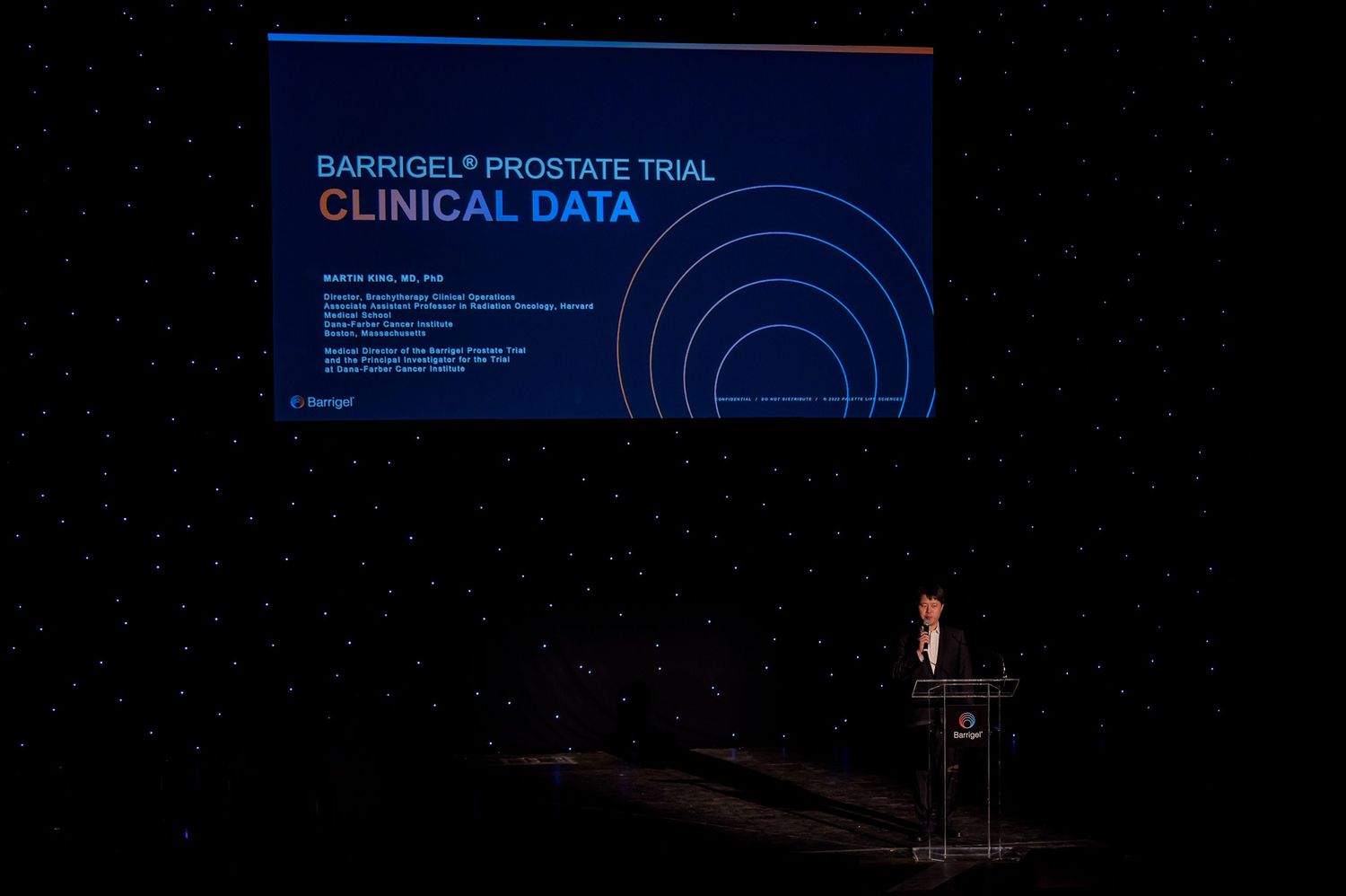 A darkened auditorium stage with a podium and blue clinical data presentation slide projected on screen.