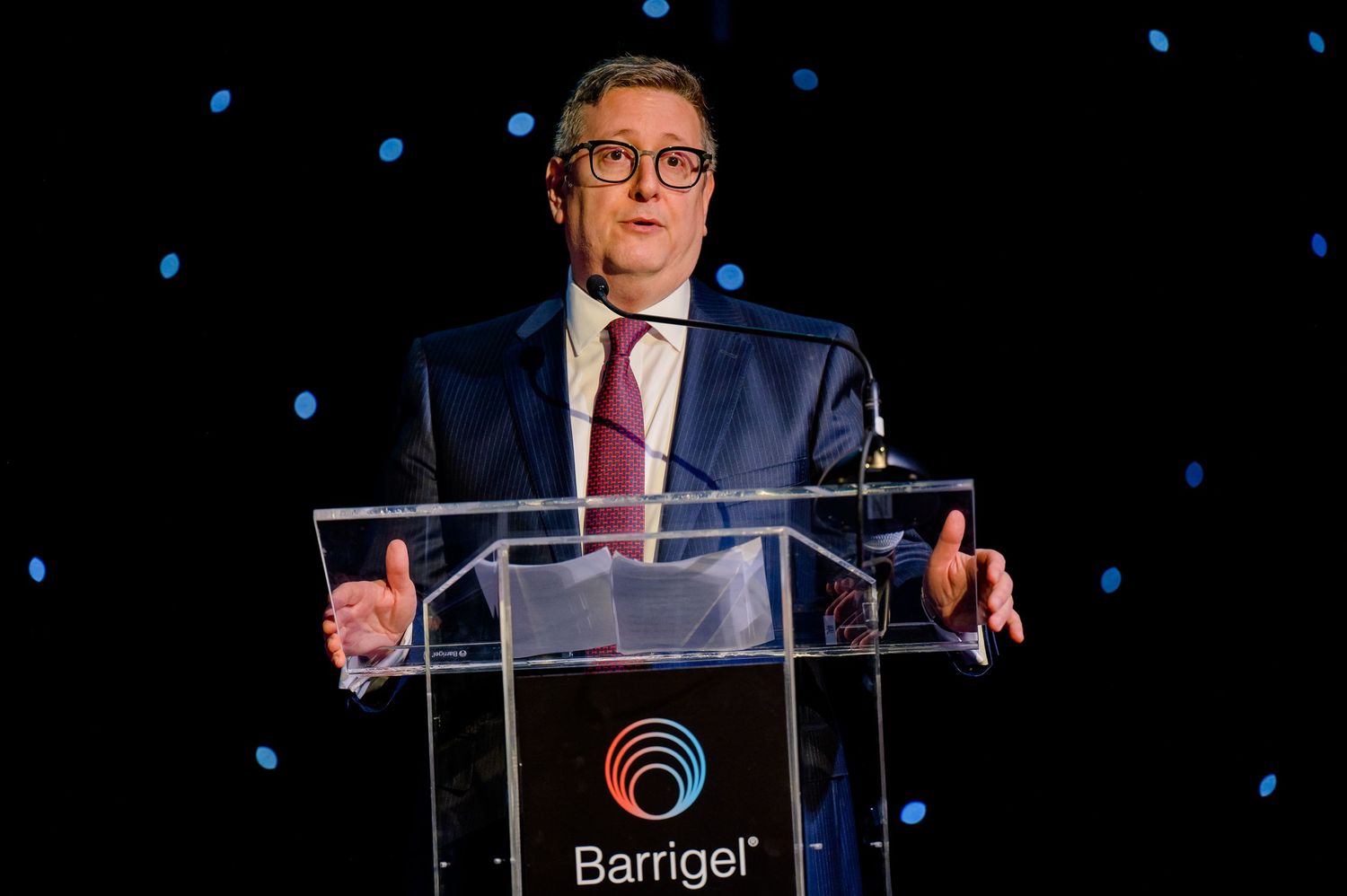 A speaker at a podium gestures with both hands while delivering remarks at a Barrigel event with blue lights in background.