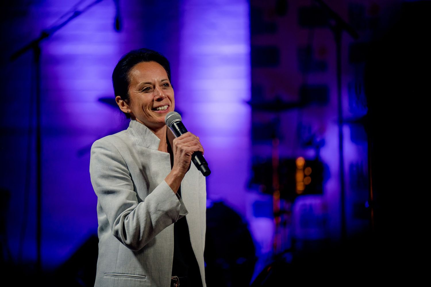 A performer wearing a white blazer sings into a microphone on stage with purple lighting in the background.