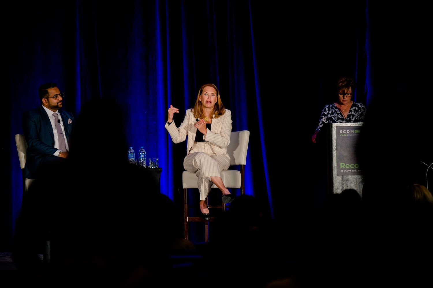Three presenters stand on a stage with blue lighting and a screen display behind them during an event panel.