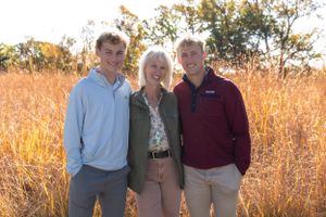 Three people pose together in casual attire against an autumn field at sunset.