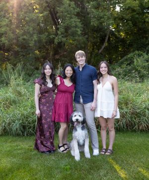 A shaggy gray and white dog poses with a group for an outdoor portrait against greenery.