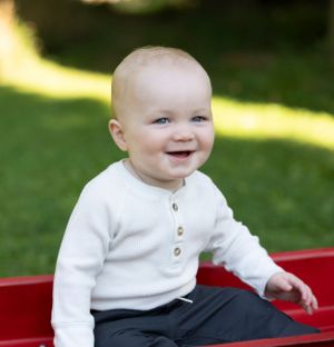 A smiling baby in a white cardigan sits on a red surface outdoors.