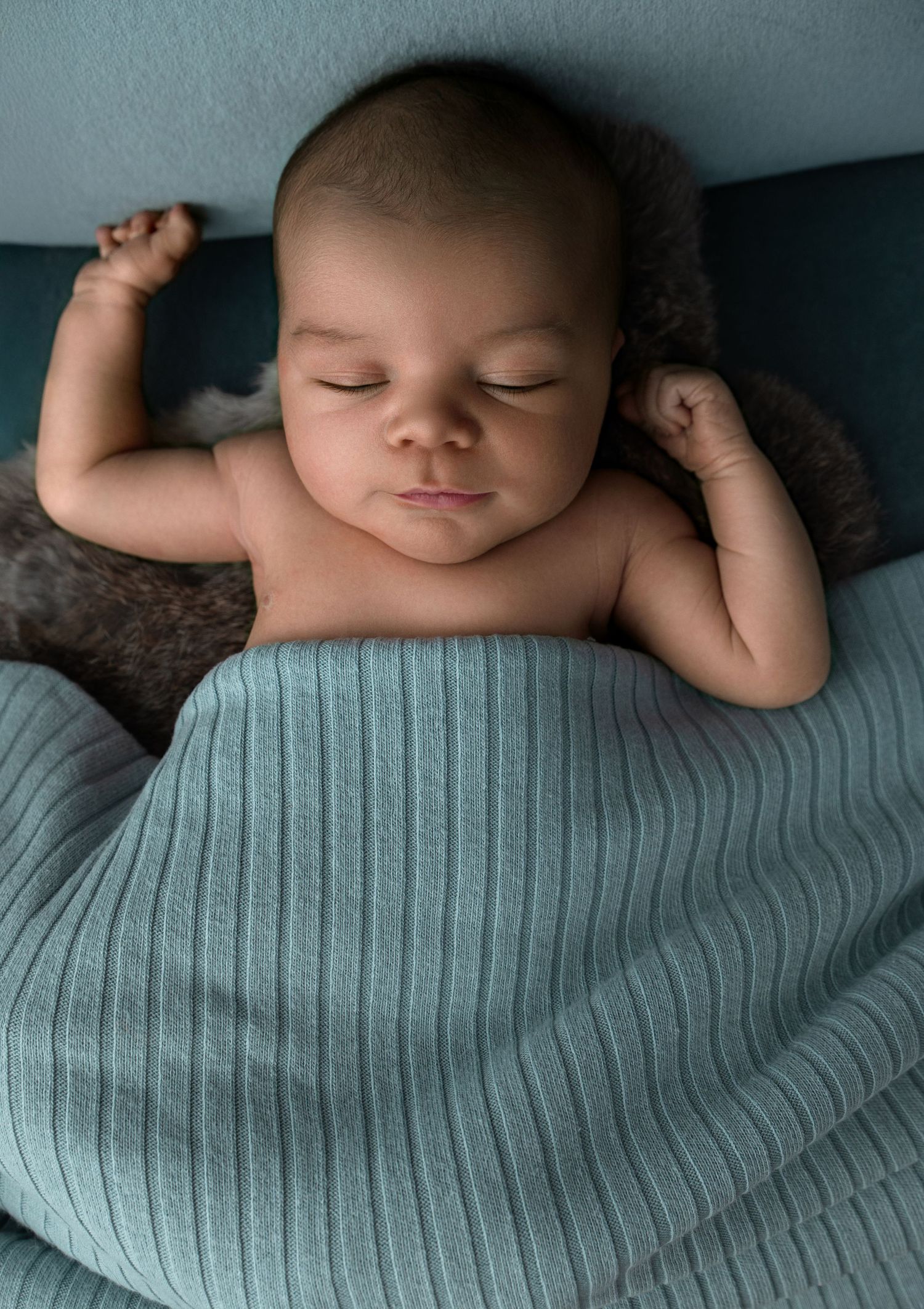 Peaceful sleeping baby lying under soft turquoise blanket with arms raised in comfortable pose.