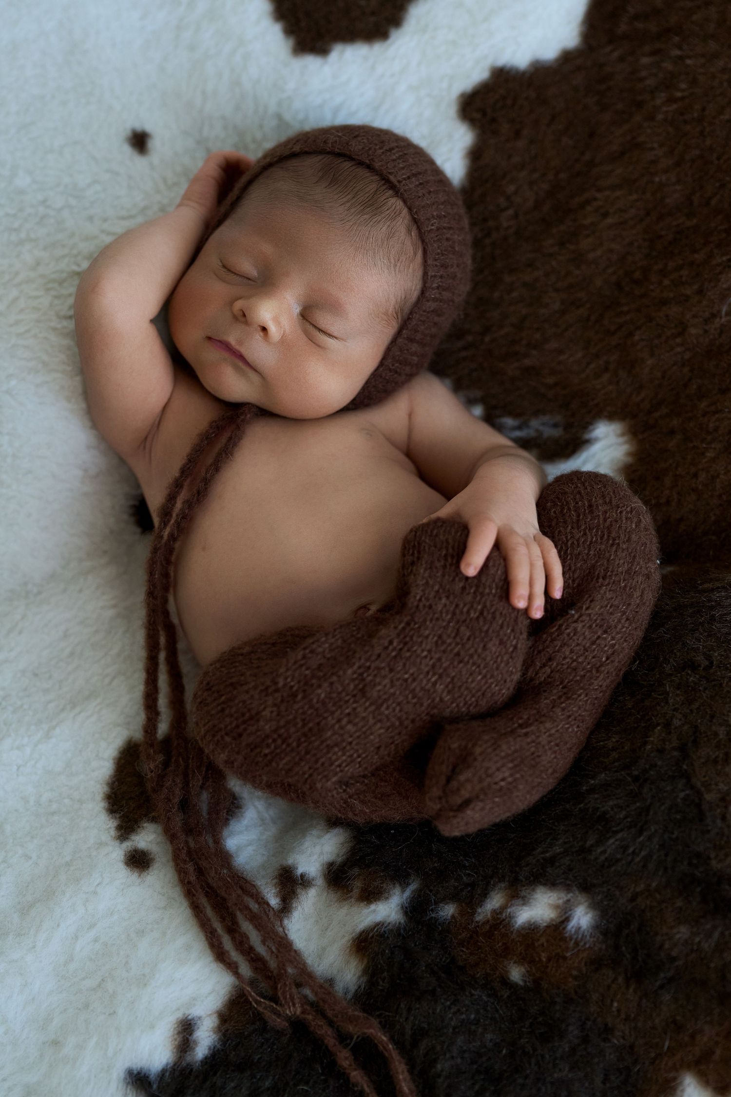 Sleeping newborn baby wearing a brown knit bonnet and wrap, posed on a cowhide rug.