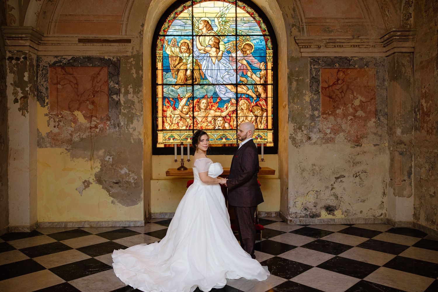 Wedding couple posing in front of stunning stained glass window in historic chapel with checkered floor.