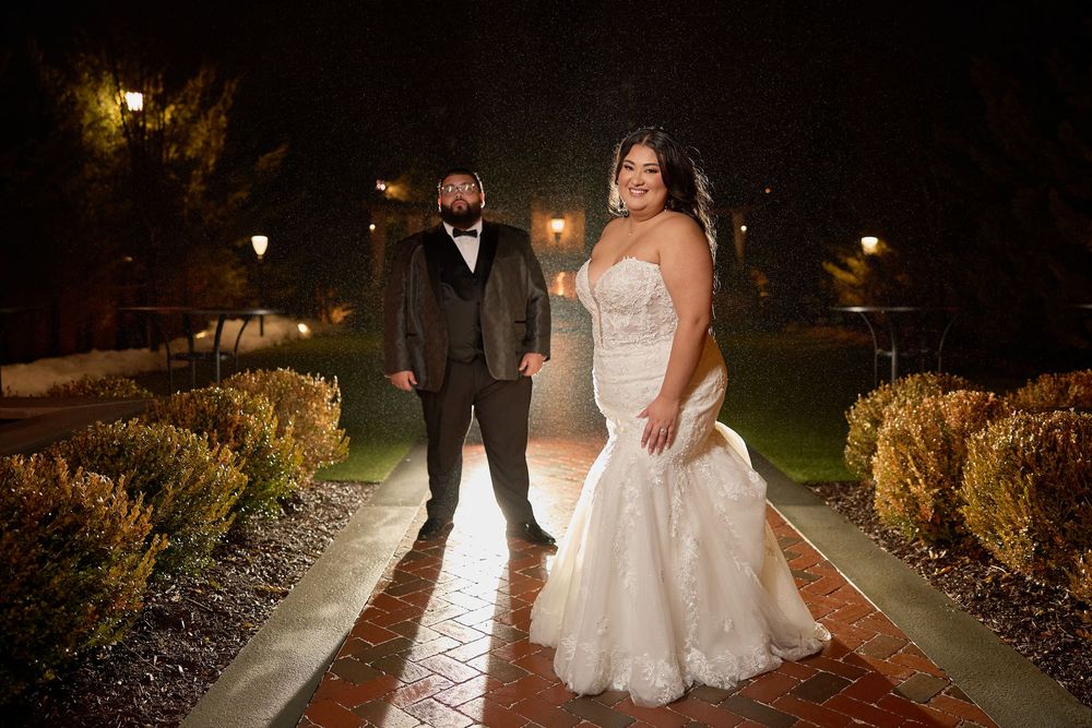 Bride in mermaid gown and groom in gray tuxedo pose on brick path at night during outdoor wedding photo.
