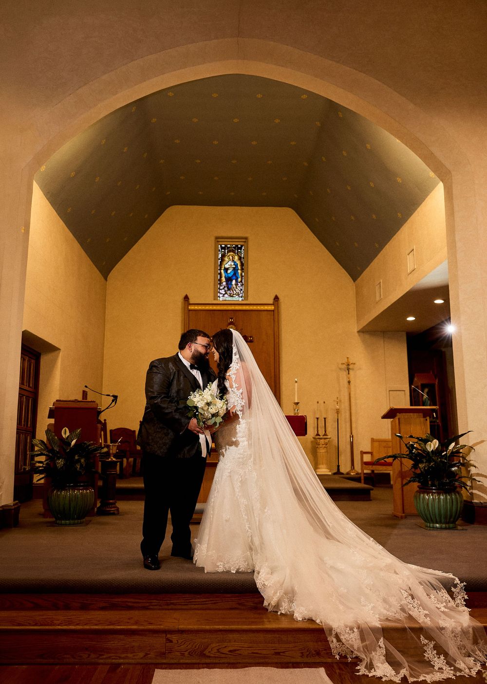 Bride and groom share a kiss at the altar of a church, with a long lace veil trailing behind the bride.