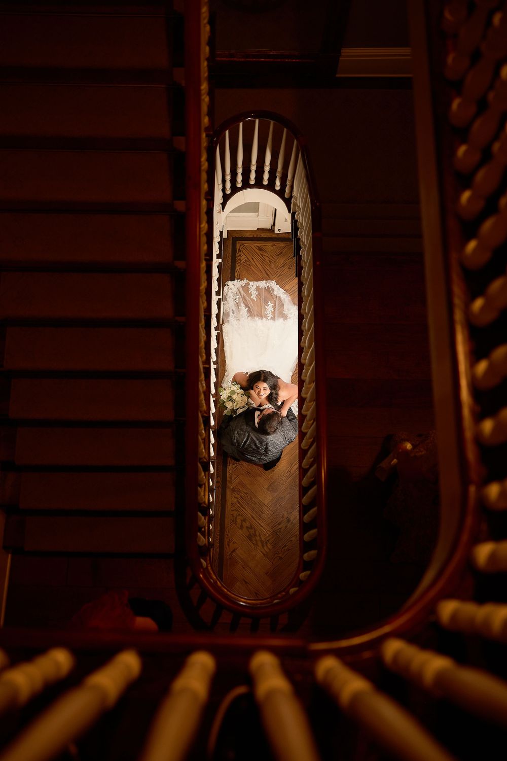 Aerial view of couple embracing in spiral staircase well, surrounded by dark wooden banisters.