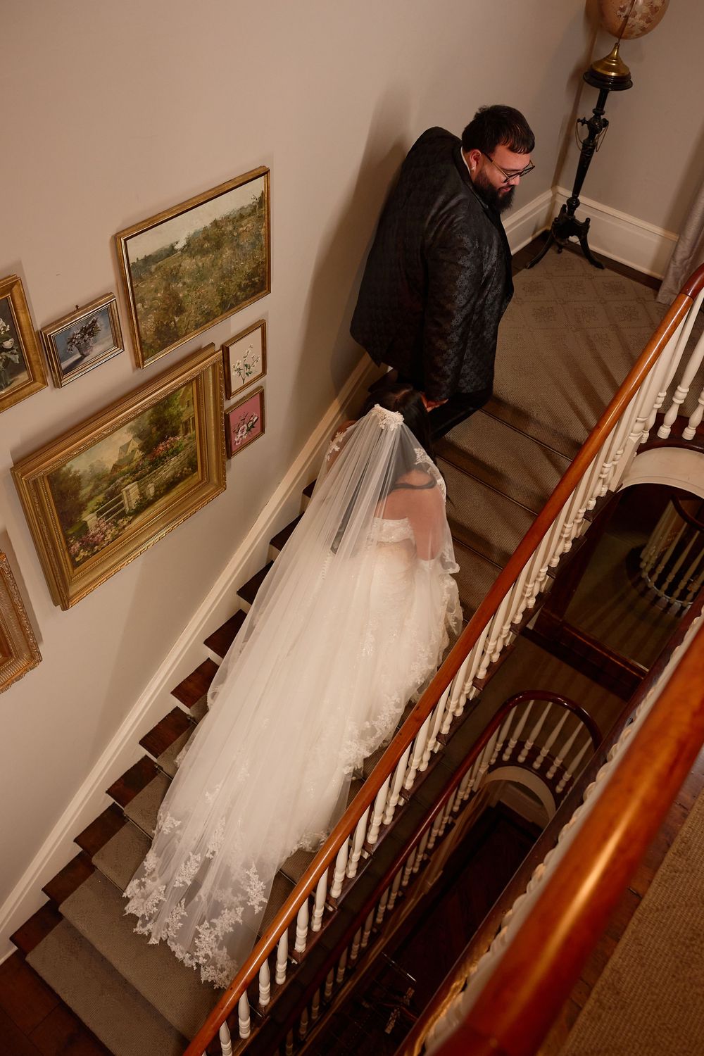 Bride in white gown and veil descends elegant wooden staircase beside suited groom, framed artwork adorning the wall.