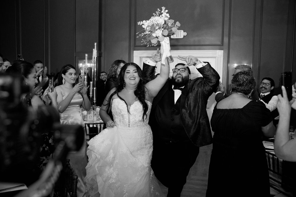 Black and white photo of a joyful bride and groom entering their wedding reception, bouquet raised high.