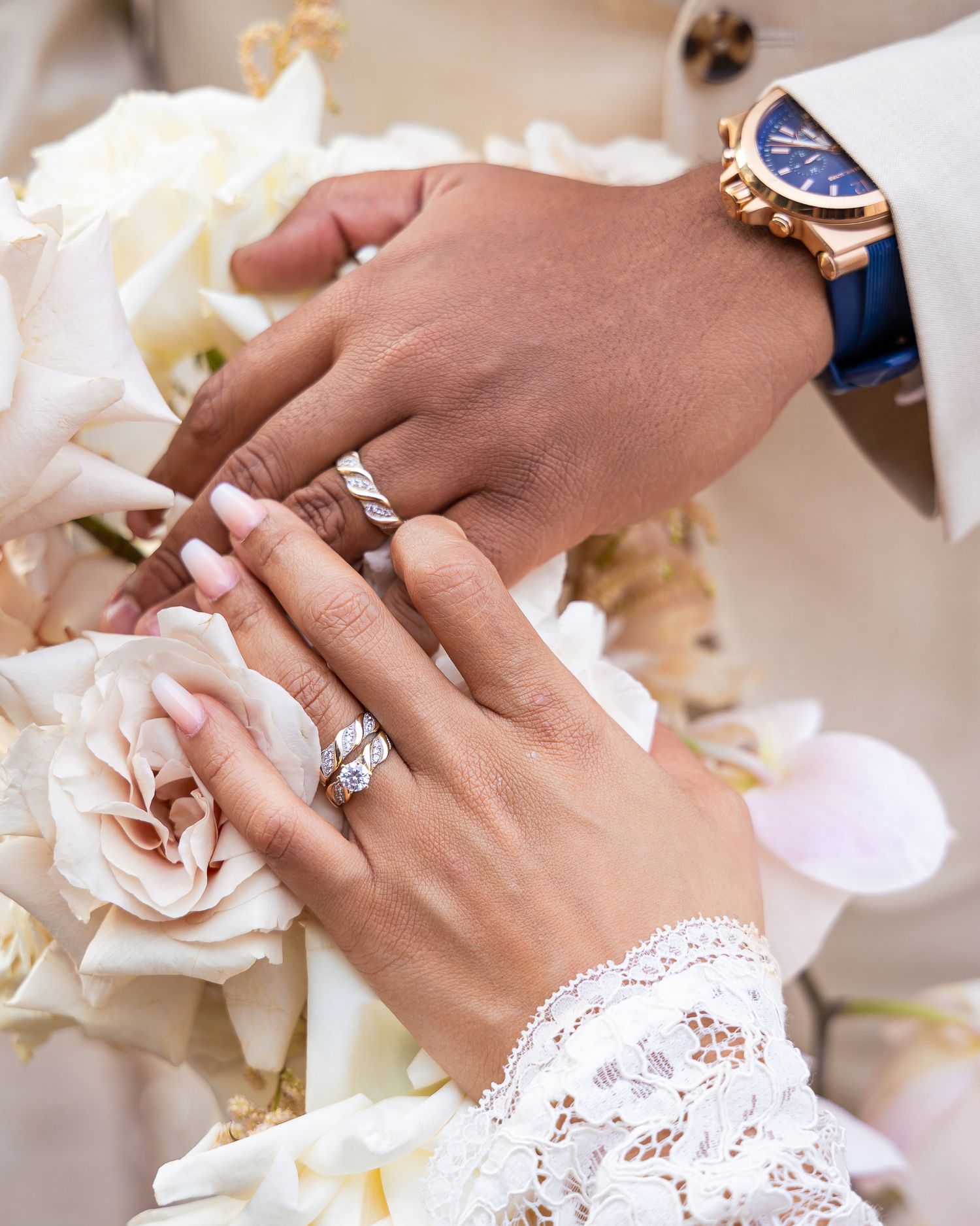 Close-up of hands wearing engagement rings with delicate rose petals and lace details in the background.