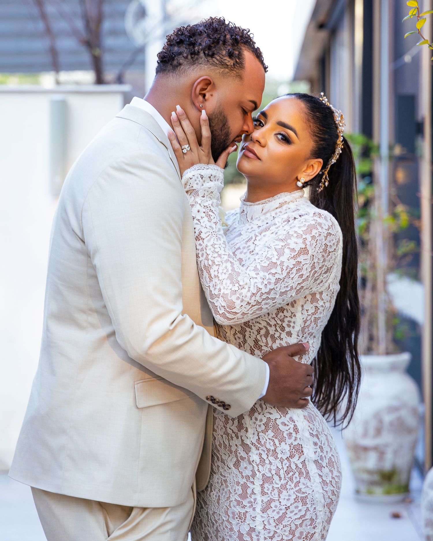 A couple in wedding attire share an intimate moment, with the bride wearing a white lace dress and groom in beige suit.