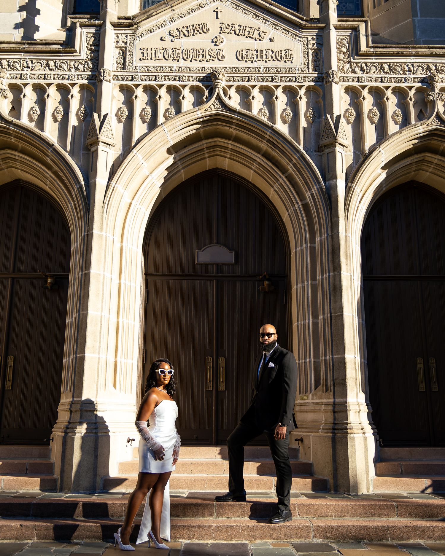 People sit and walk under ornate Gothic archways at a historic university building entrance in afternoon sunlight.