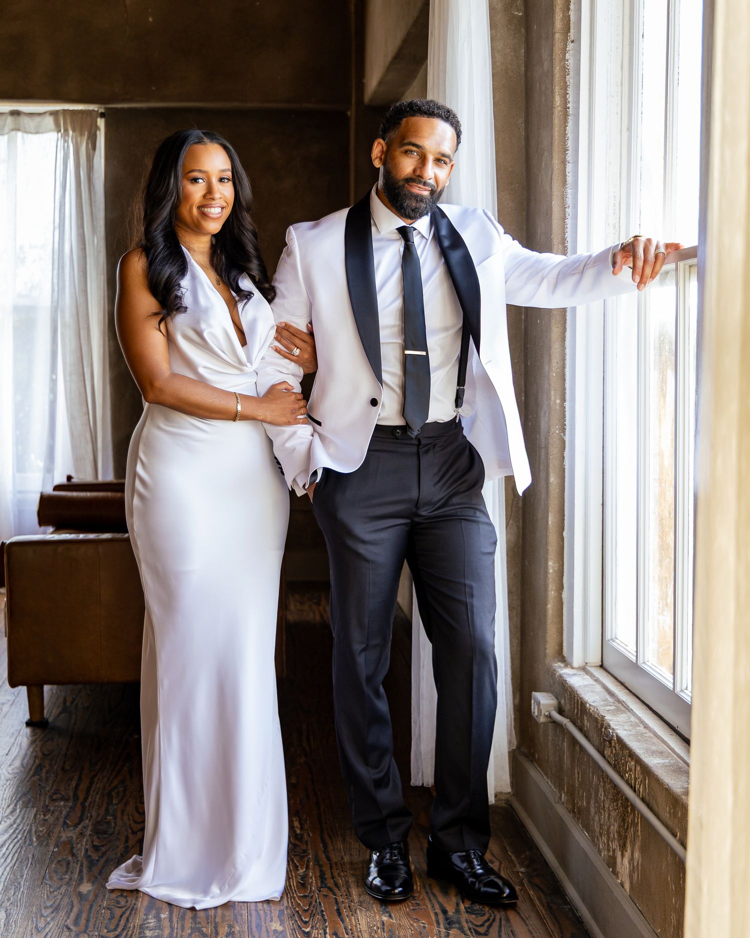A couple in formal attire poses elegantly by a window, with the woman in a white gown and man in a light blazer and tie.