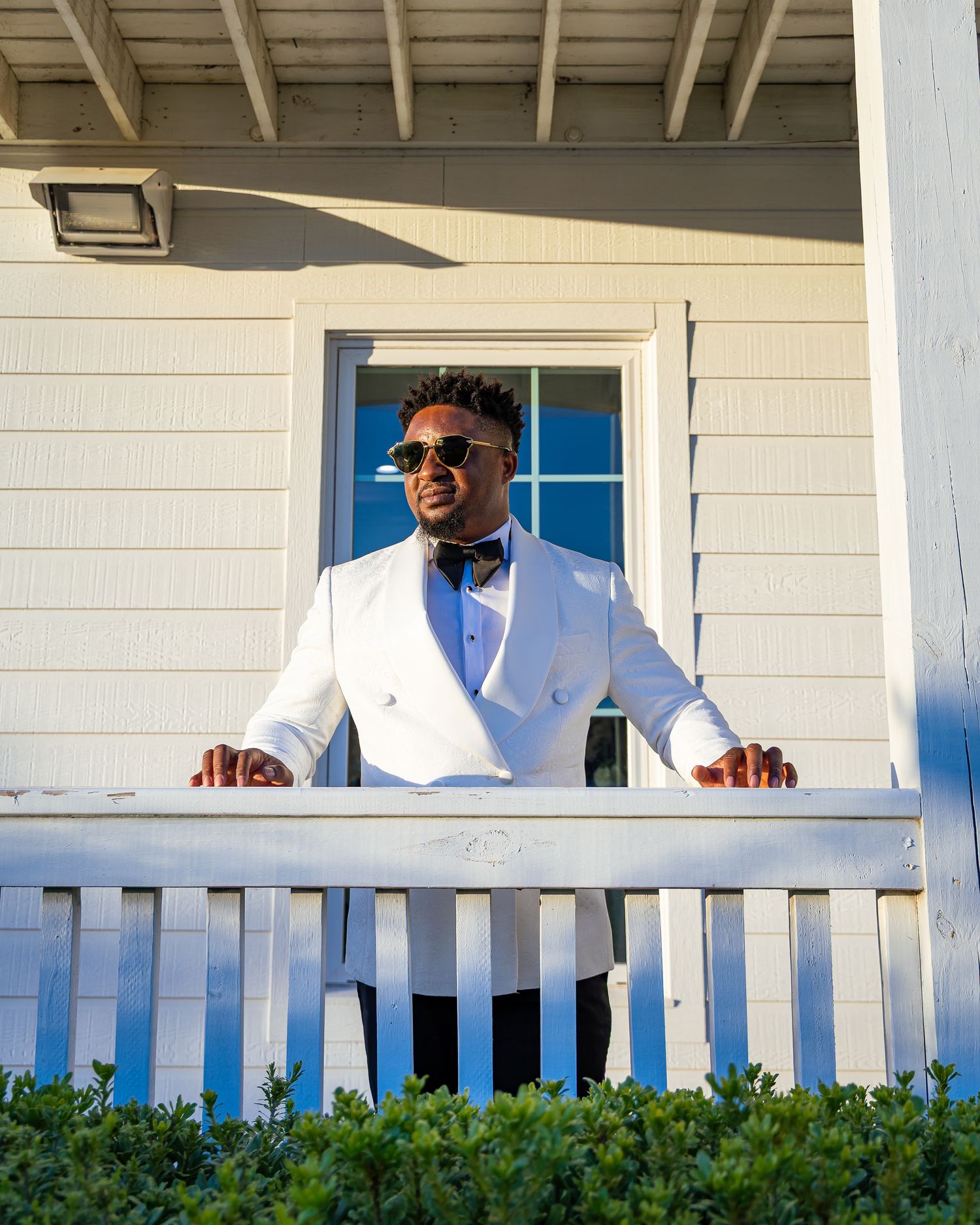 Person wearing a white shirt and sunglasses smiles while standing on a porch with white railing and greenery.