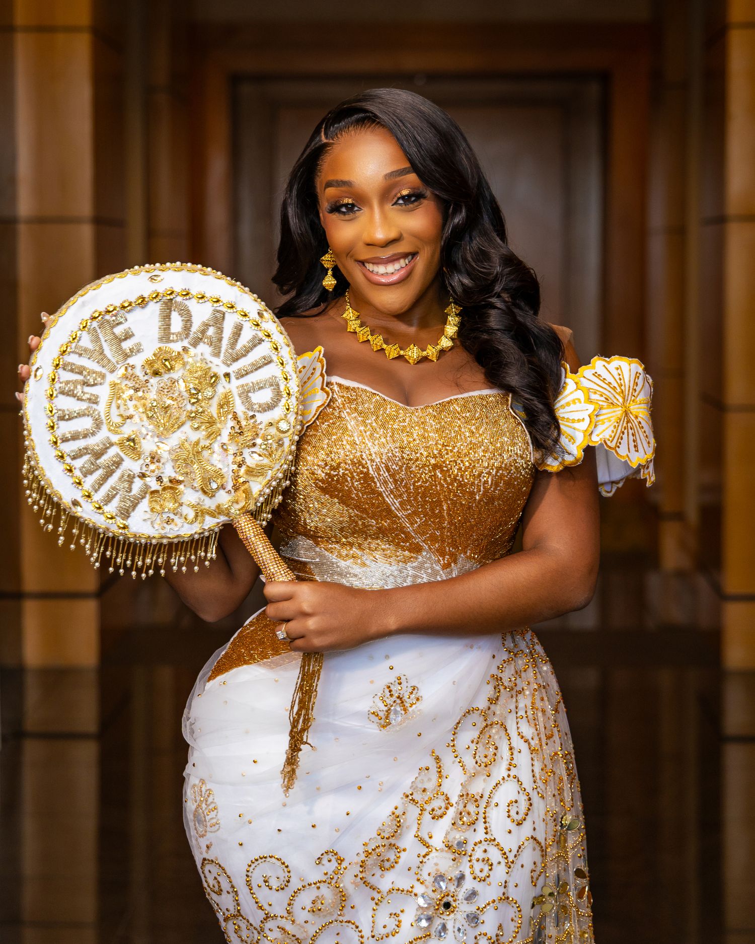 A person in a sparkling gold and white dress holds a decorative sign with 'DAD' written in pearls.