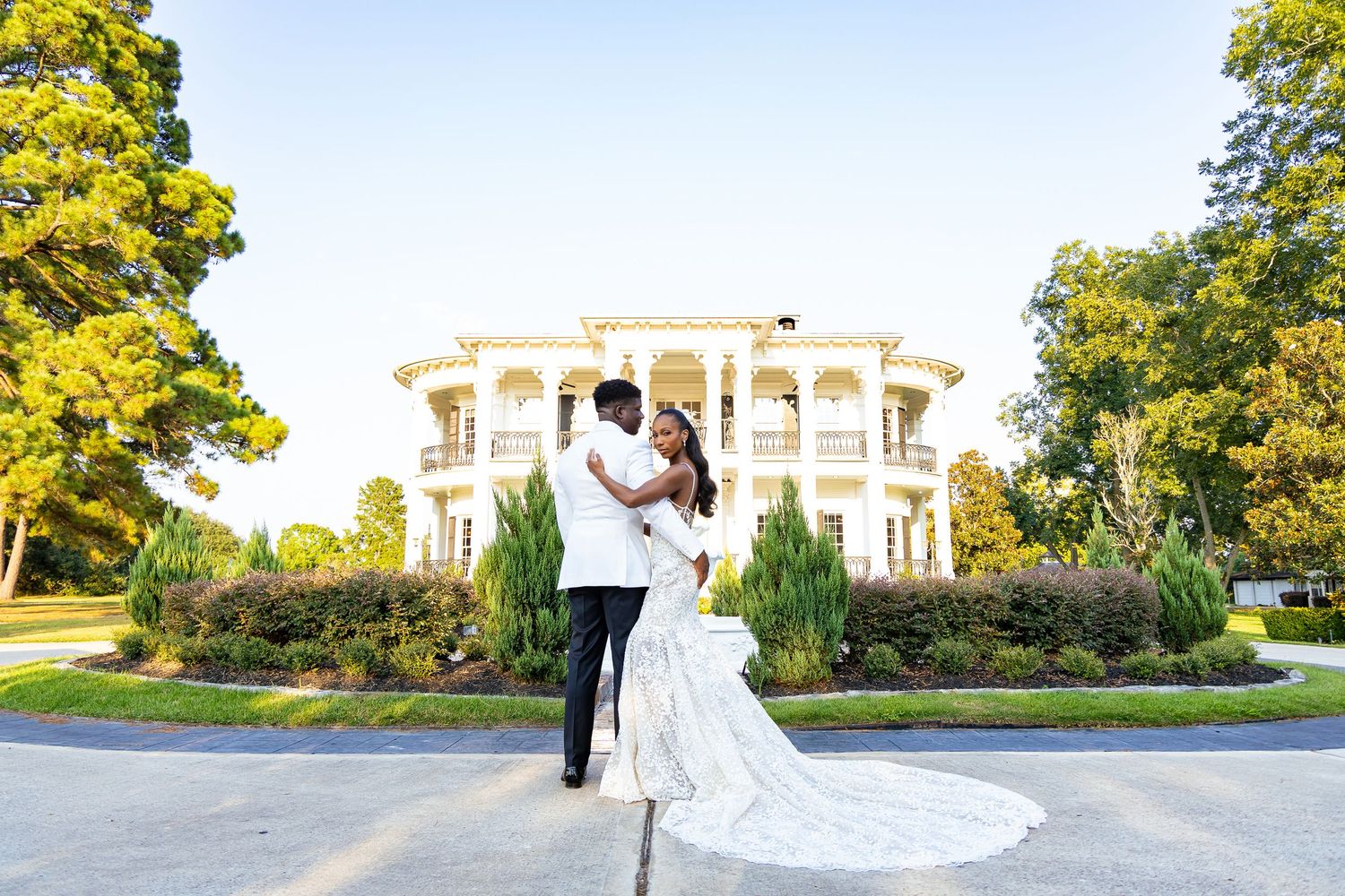 Wedding couple walks through a lush garden path with sunlight filtering through trees during an outdoor photo session.
