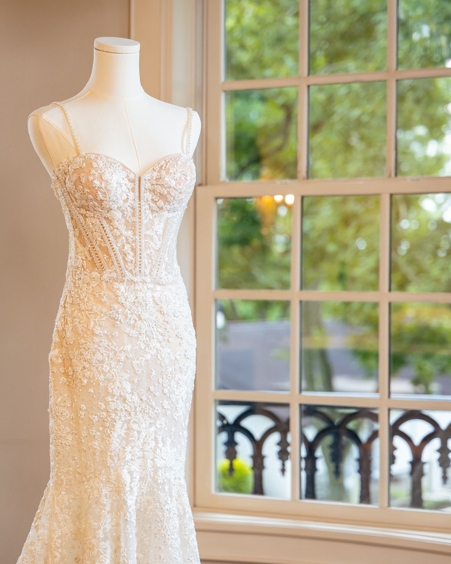 White lace wedding dress with delicate beading details displayed against window with natural lighting.