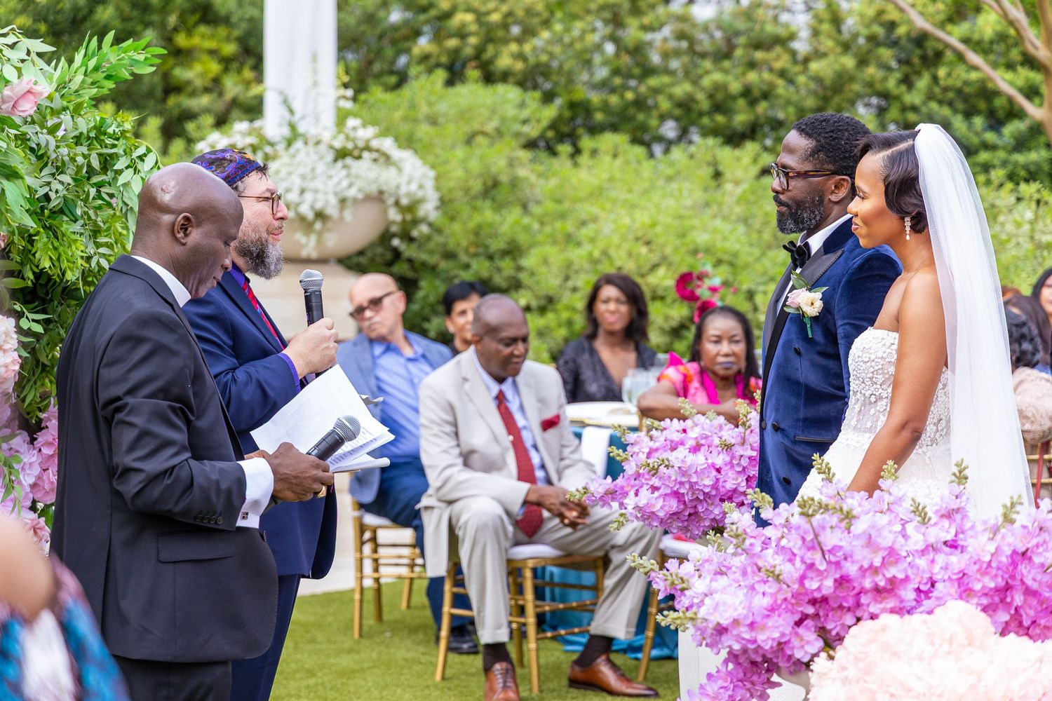 Outdoor garden wedding ceremony with guests seated in white chairs surrounded by lush greenery and pink flowers.