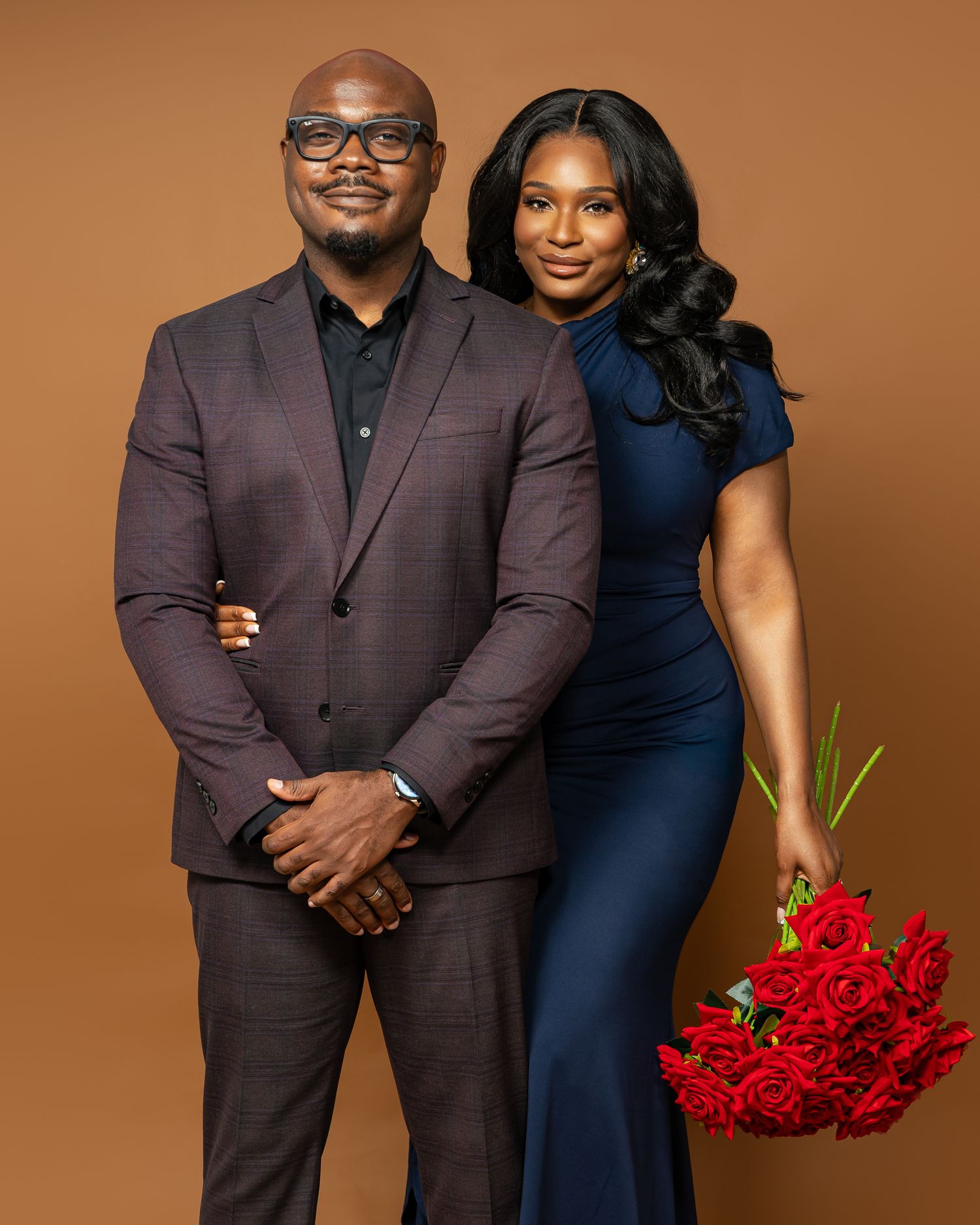 A couple in formal attire poses against a brown backdrop, with the woman holding red roses in her navy blue dress.