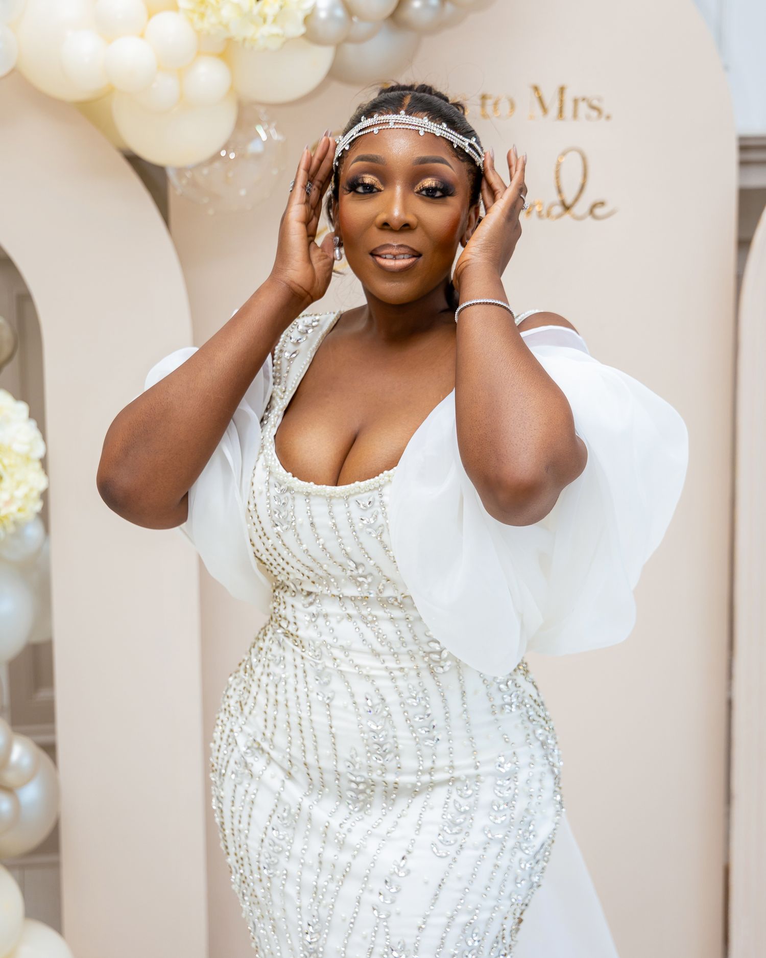 A bride in a white beaded wedding dress poses with white balloons in an elegant indoor setting.