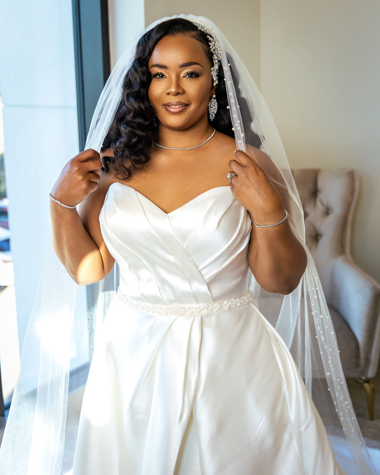 Bride in an elegant white satin wedding dress with a plunging neckline poses against a bright window backdrop.