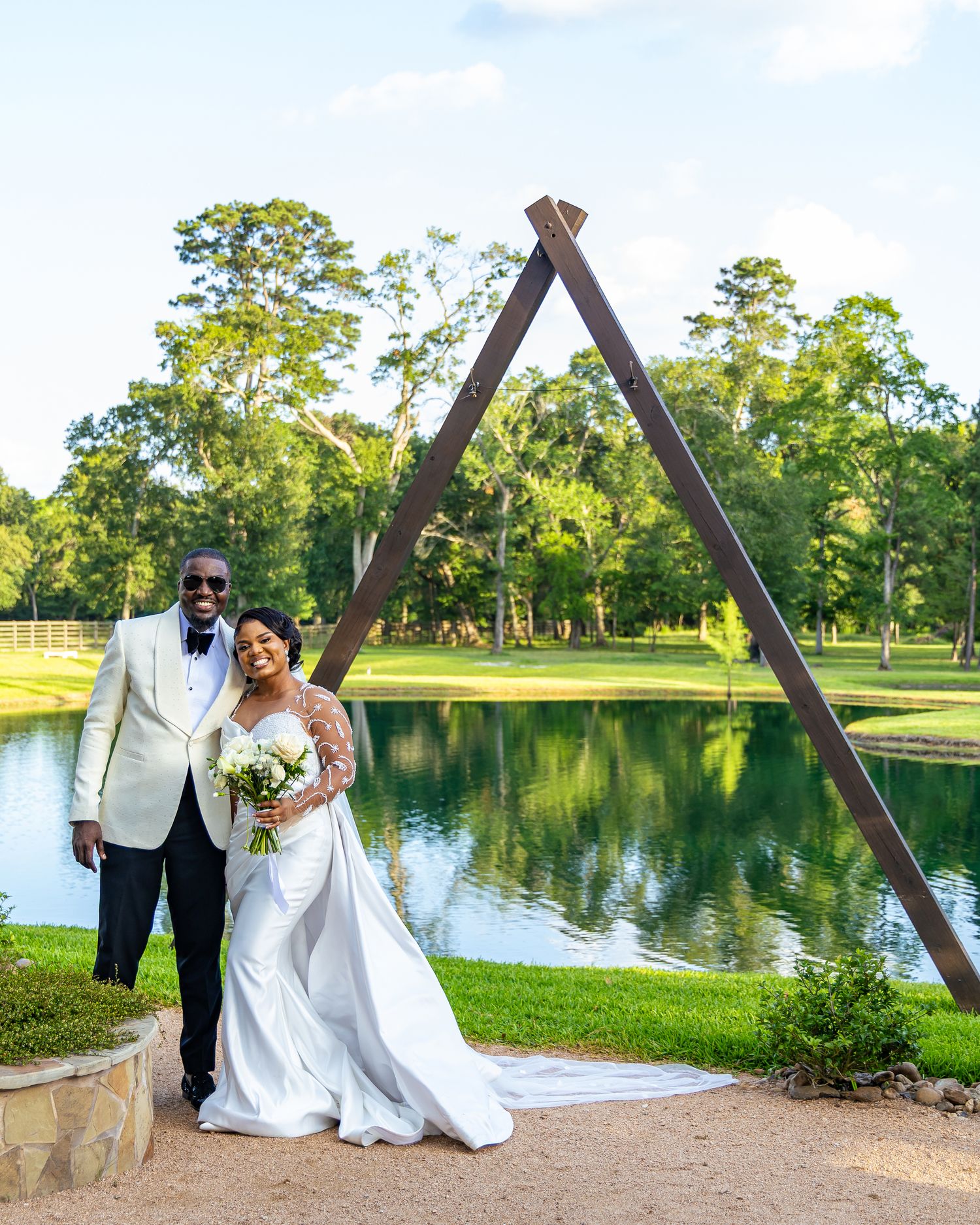 A wedding couple shares a romantic moment by a serene lake surrounded by lush greenery in a scenic park setting.