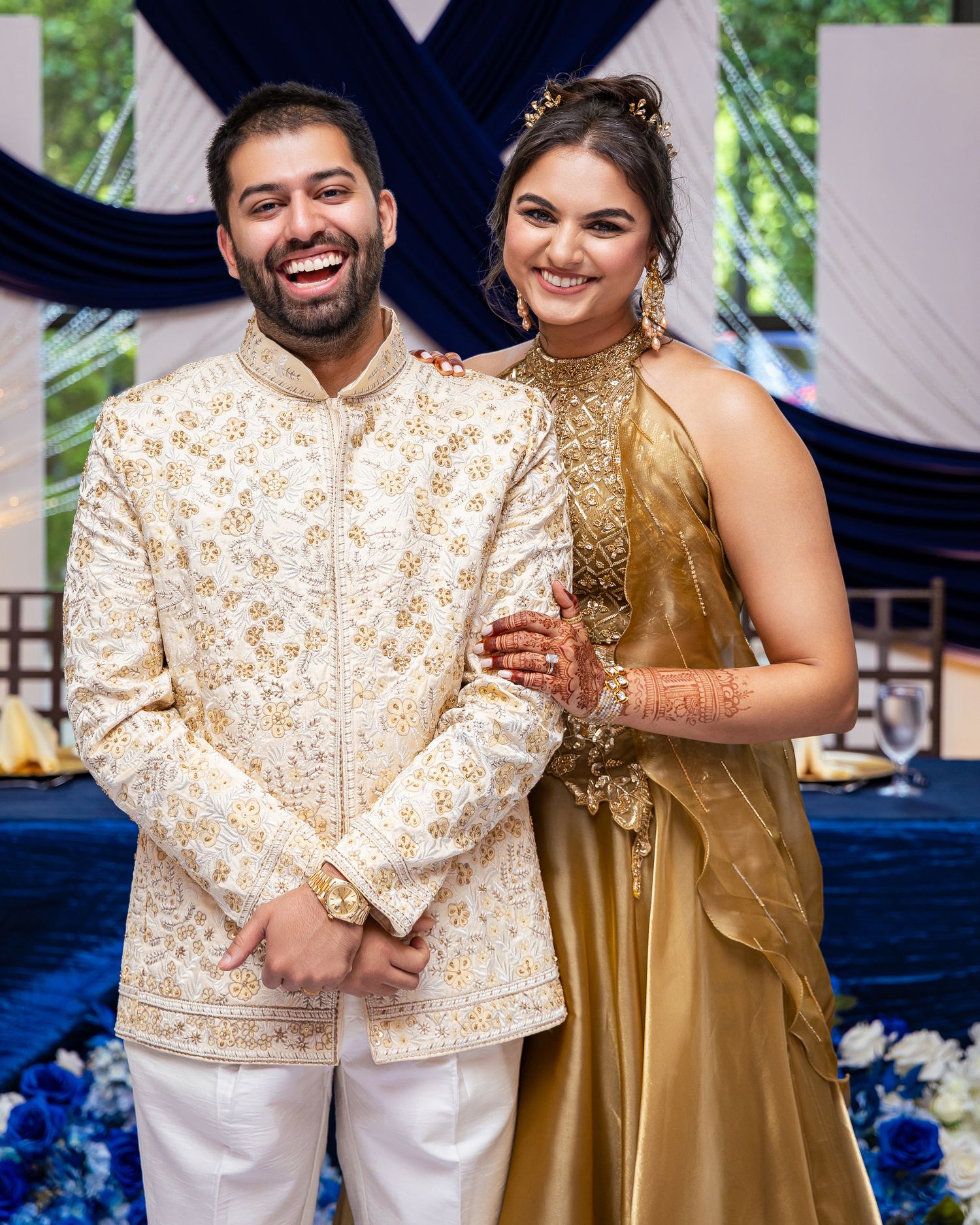 A couple in traditional Indian wedding attire pose together, with the groom in white and gold and bride in gold.