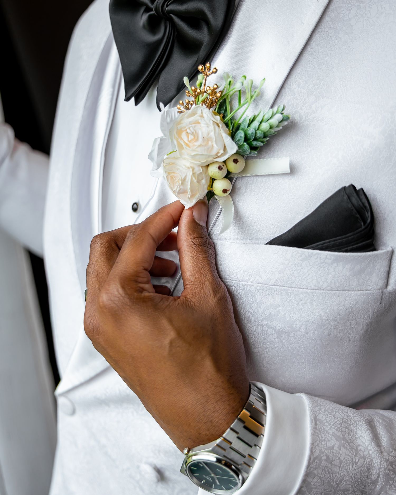 A hand adjusts an elegant white rose and succulent boutonniere on a white tuxedo jacket with black bow tie.