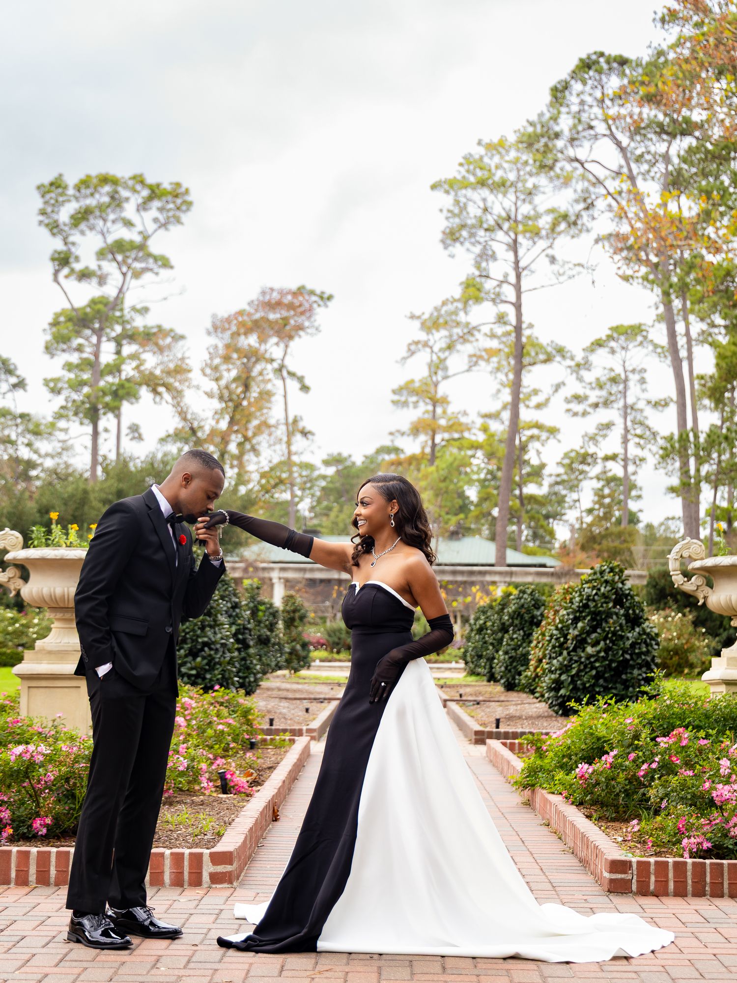 A couple shares a romantic moment in formal attire on a brick patio surrounded by greenery and pine trees.