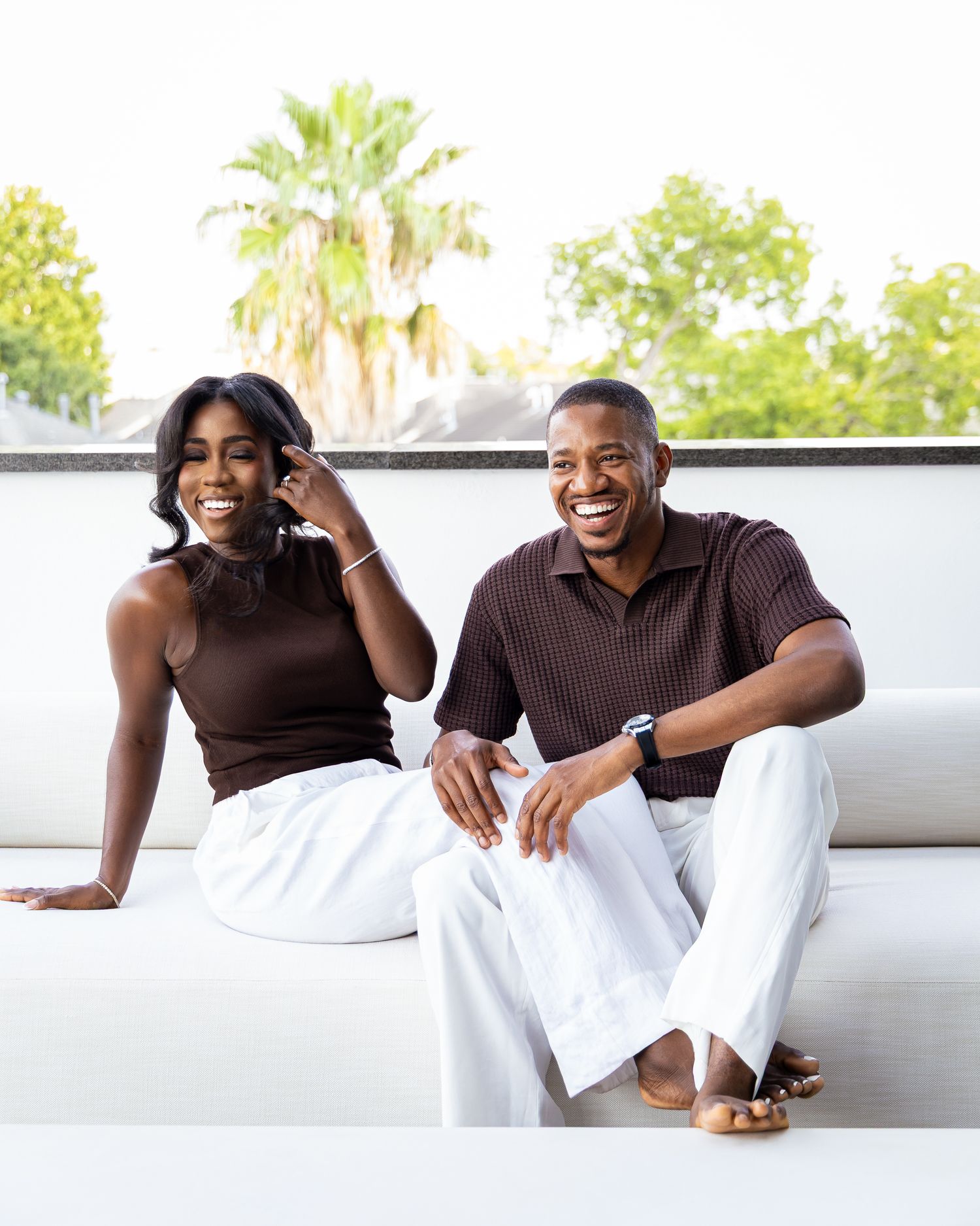 Two people in brown tops and white pants sit casually together on an outdoor couch with palm trees visible in background.
