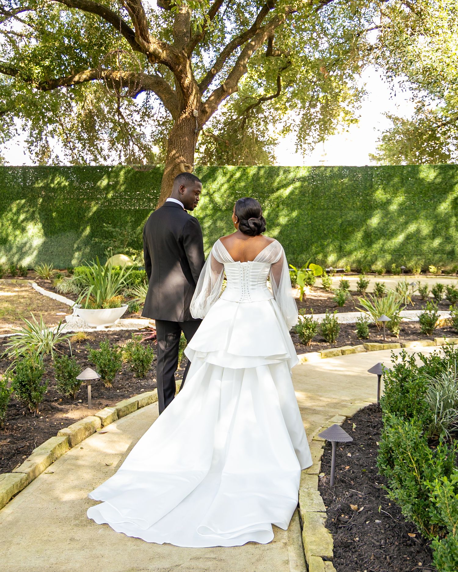 A bride and groom walk along a garden path surrounded by lush greenery and trees on their wedding day.