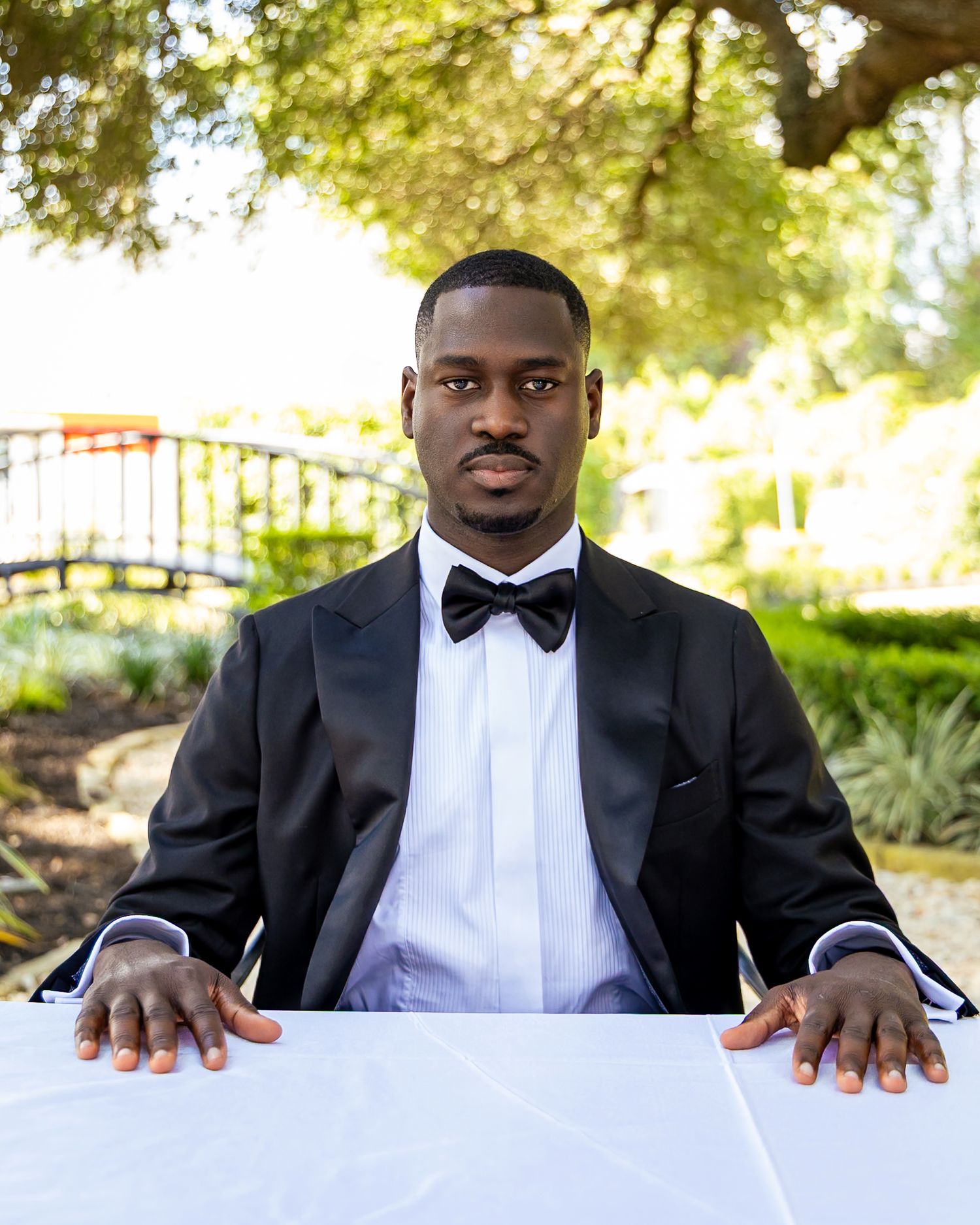 A groom in a classic black tuxedo sits outdoors during a wedding photoshoot in a garden setting.