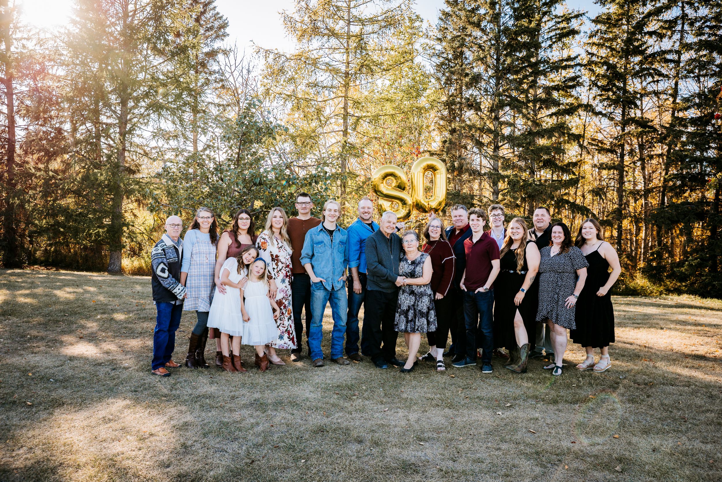 Large family group celebrating a 30th anniversary outdoors with gold balloon numbers in forest.