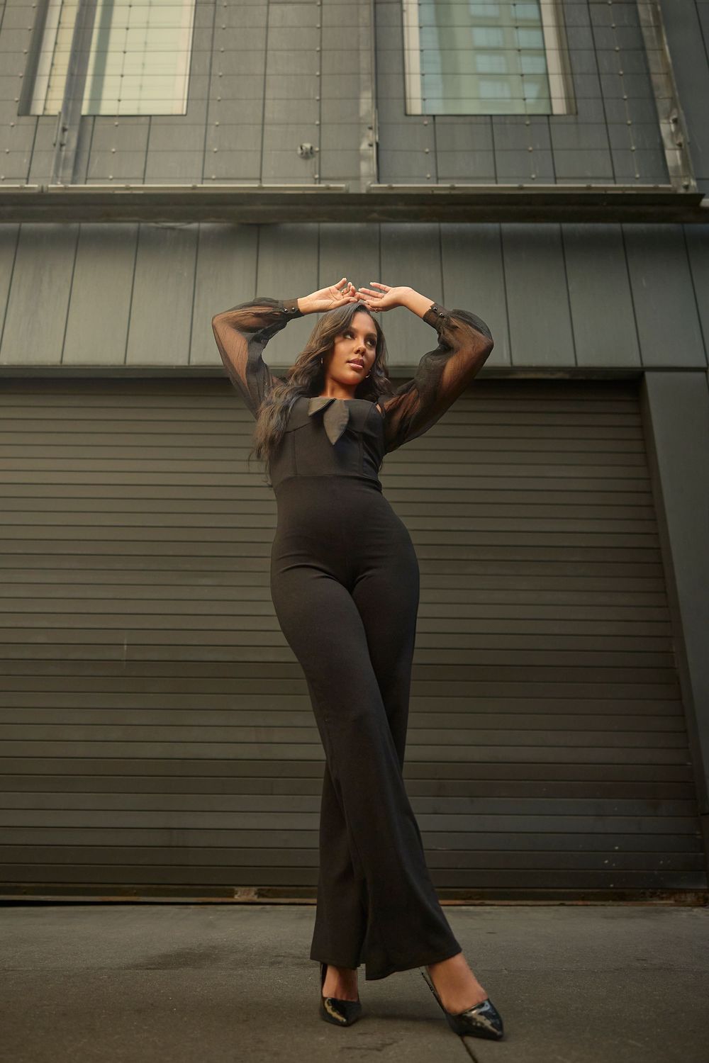 A series of fashion poses against a gray industrial wall showcasing a fitted black dress and heels.