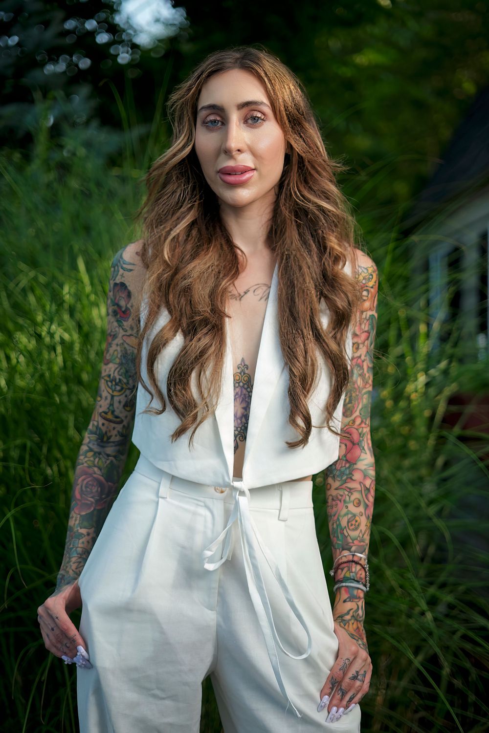 Fashion portrait of tattooed woman in white jumpsuit against garden background.