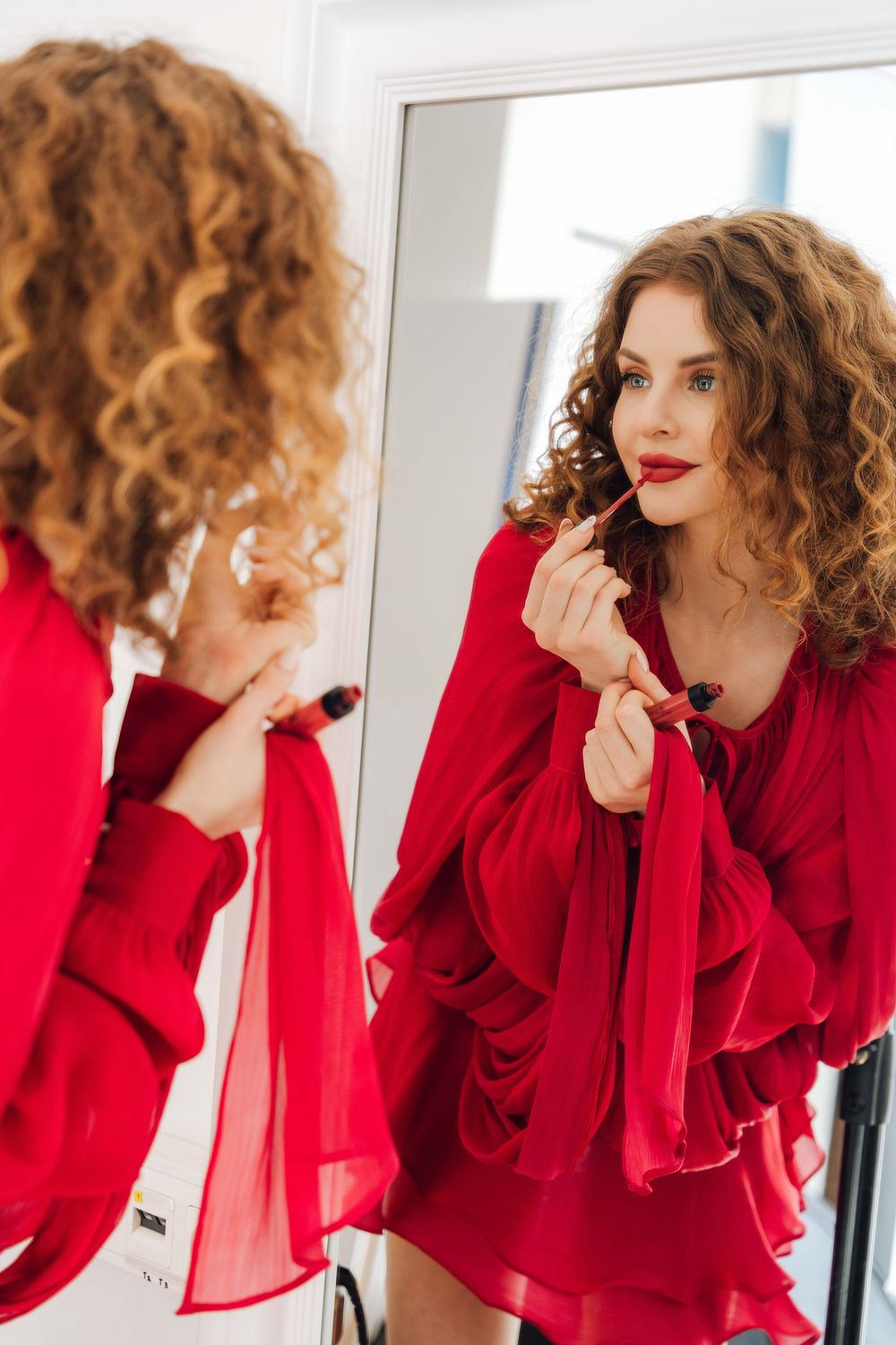 Model in flowing red dress applies lipstick while looking in mirror.
