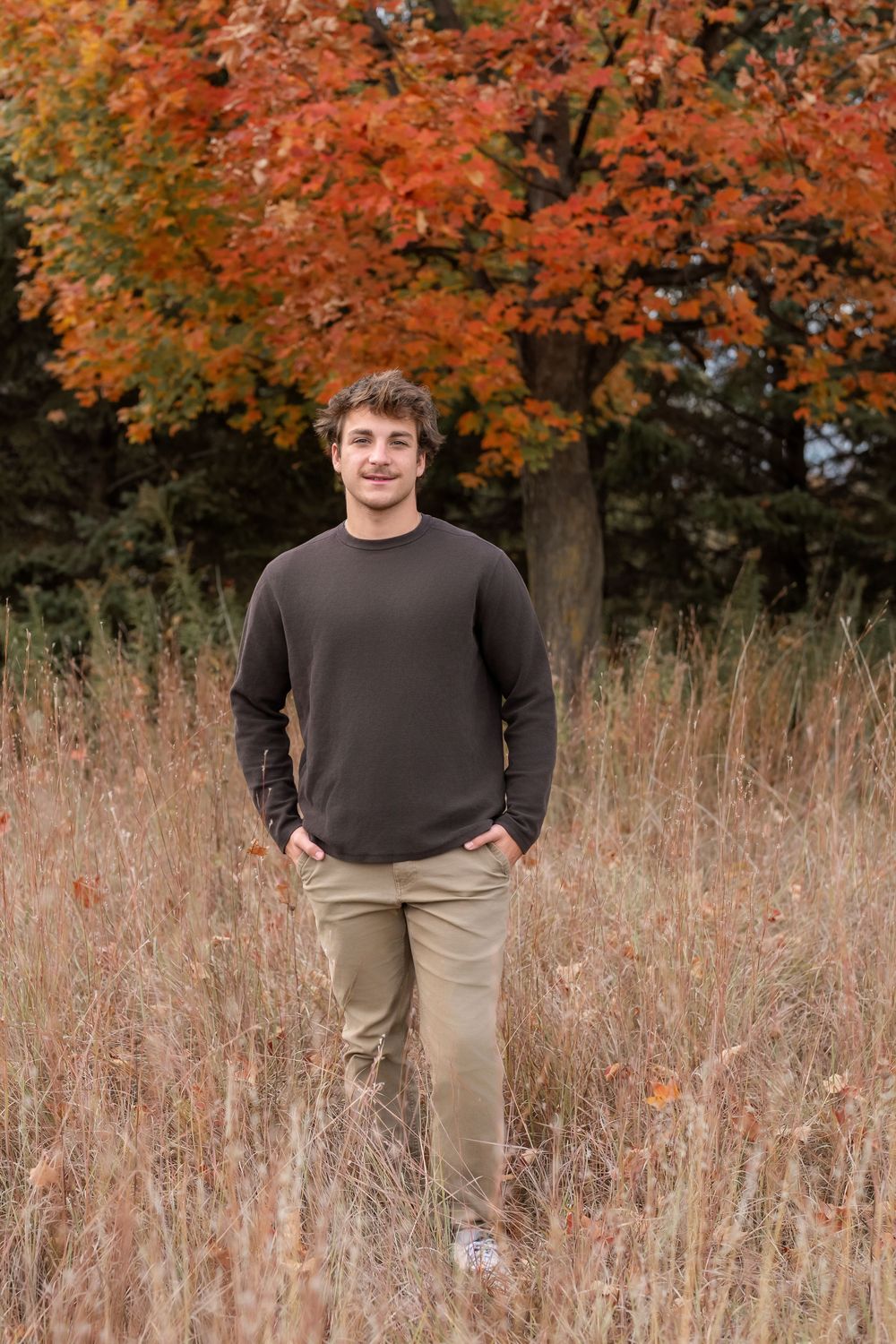 Young man in brown sweater and khaki pants smiling in autumn field with colorful orange foliage trees.