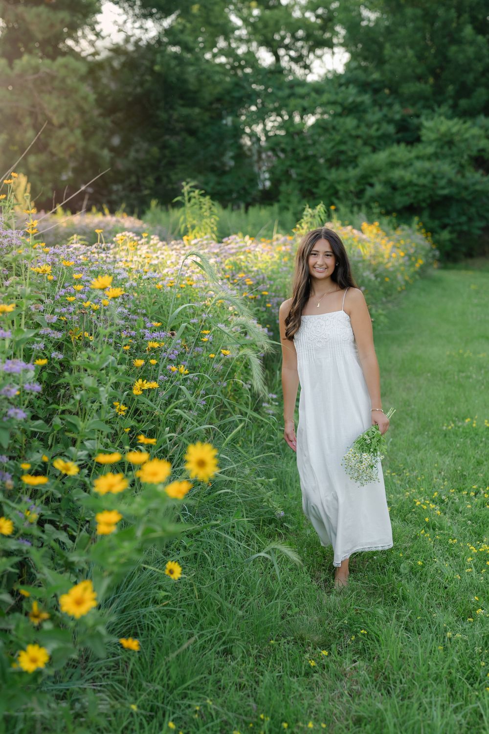 Young woman in white dress walks through wildflower meadow holding bouquet, surrounded by yellow and purple blooms.