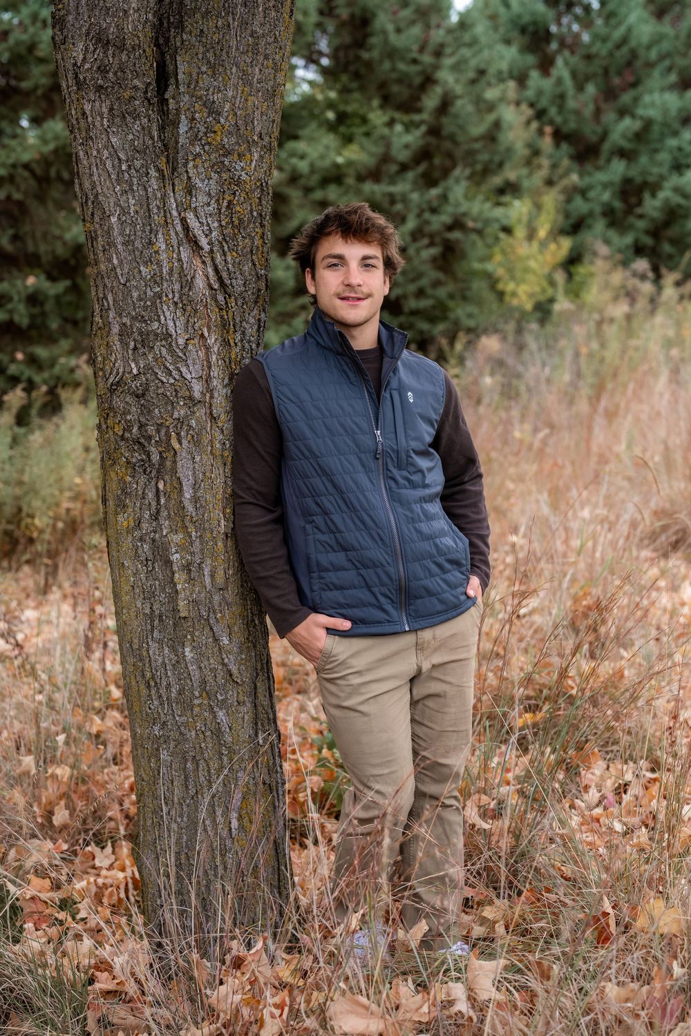 Young man in blue vest and khakis leaning against a tree in an autumn forest setting.