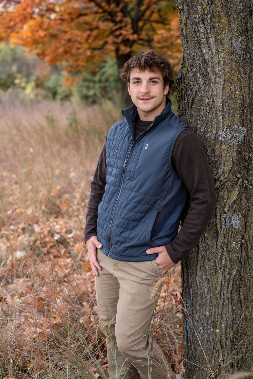 Young man in blue vest leaning against tree in autumn forest setting with colorful fall foliage.