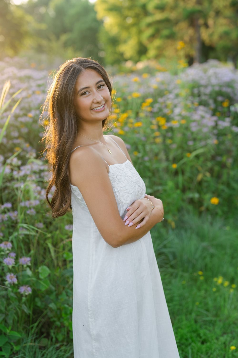 Young woman in white sundress smiling in a wildflower meadow with golden sunlight and purple and yellow blooms.