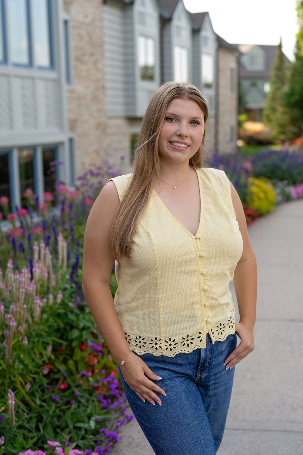 Young woman with long blonde hair smiles in yellow sleeveless top and jeans, posing by colorful garden flowers.