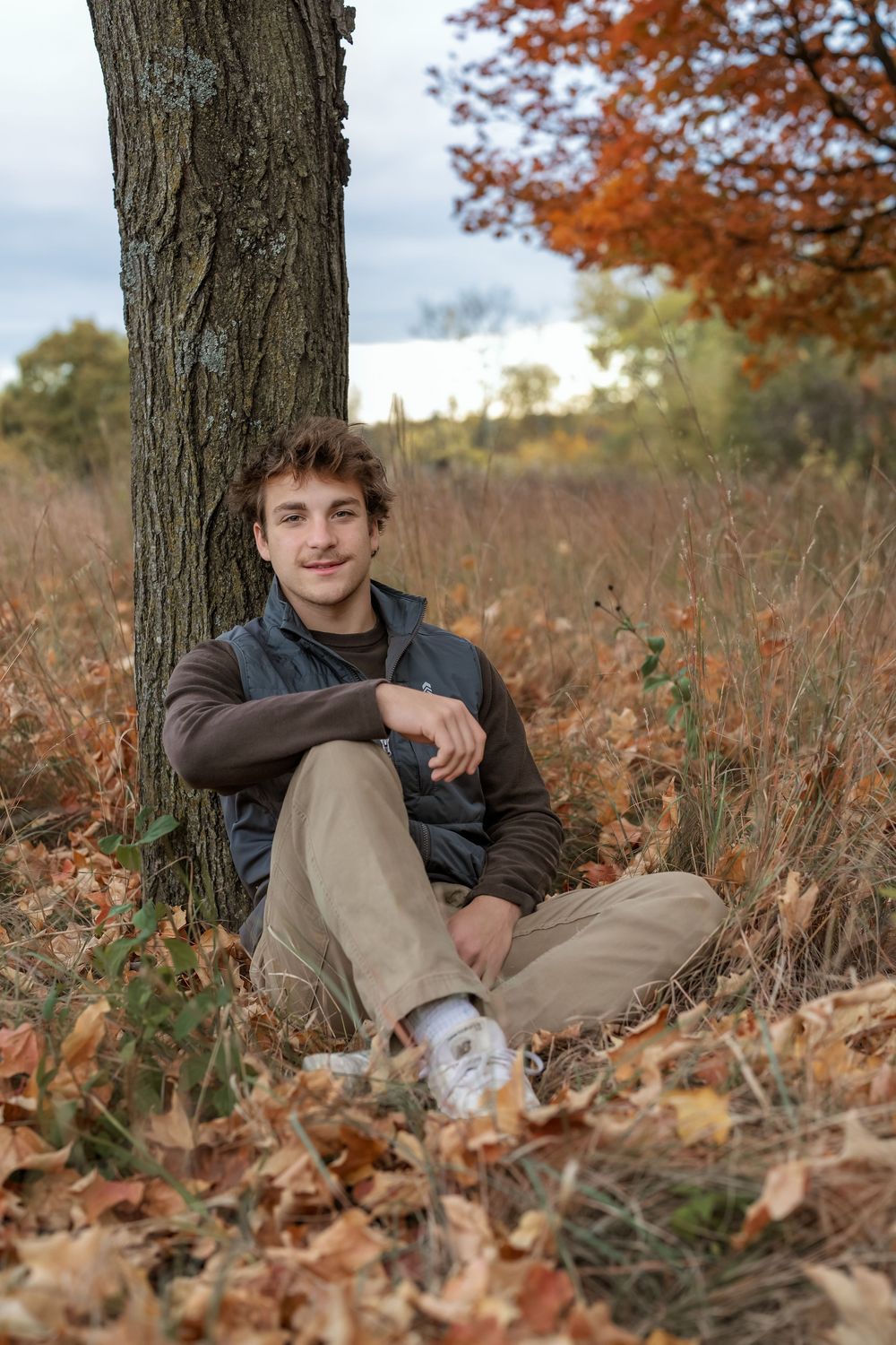 Young man sitting against a tree in autumn field, wearing vest and khakis, smiling among fallen leaves.